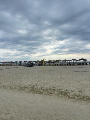Ein Strand mit Sonnenschirmen, Liegestühlen, einem Volleyballnetz und einem bewölkten Himmel.
