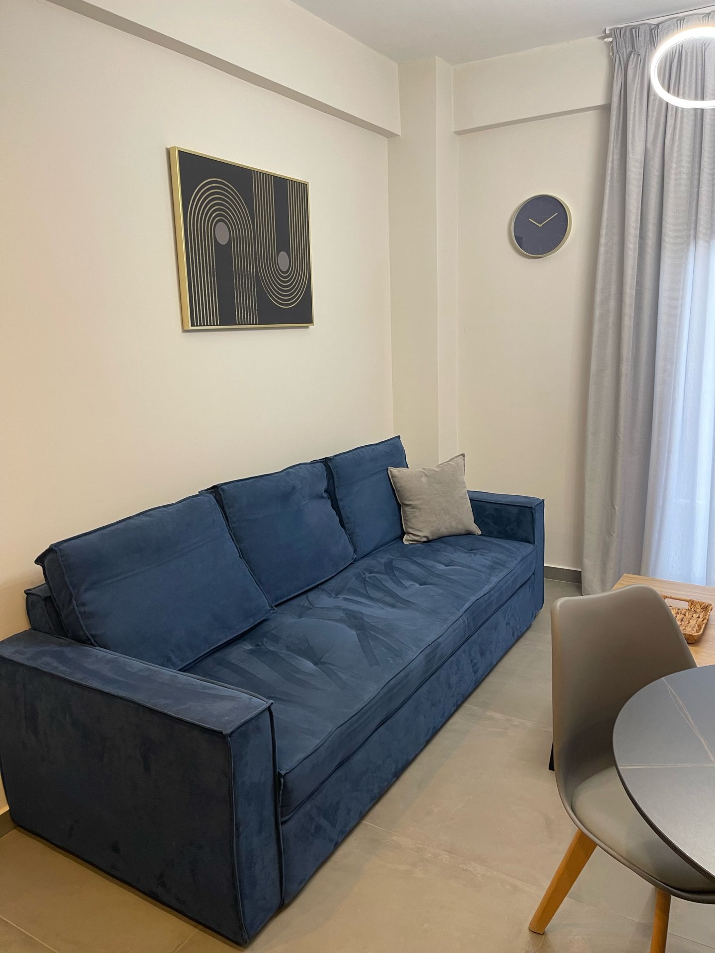 Un canapé bleu avec un coussin, une table ronde avec une chaise, une horloge murale et une œuvre d’art abstraite encadrée.