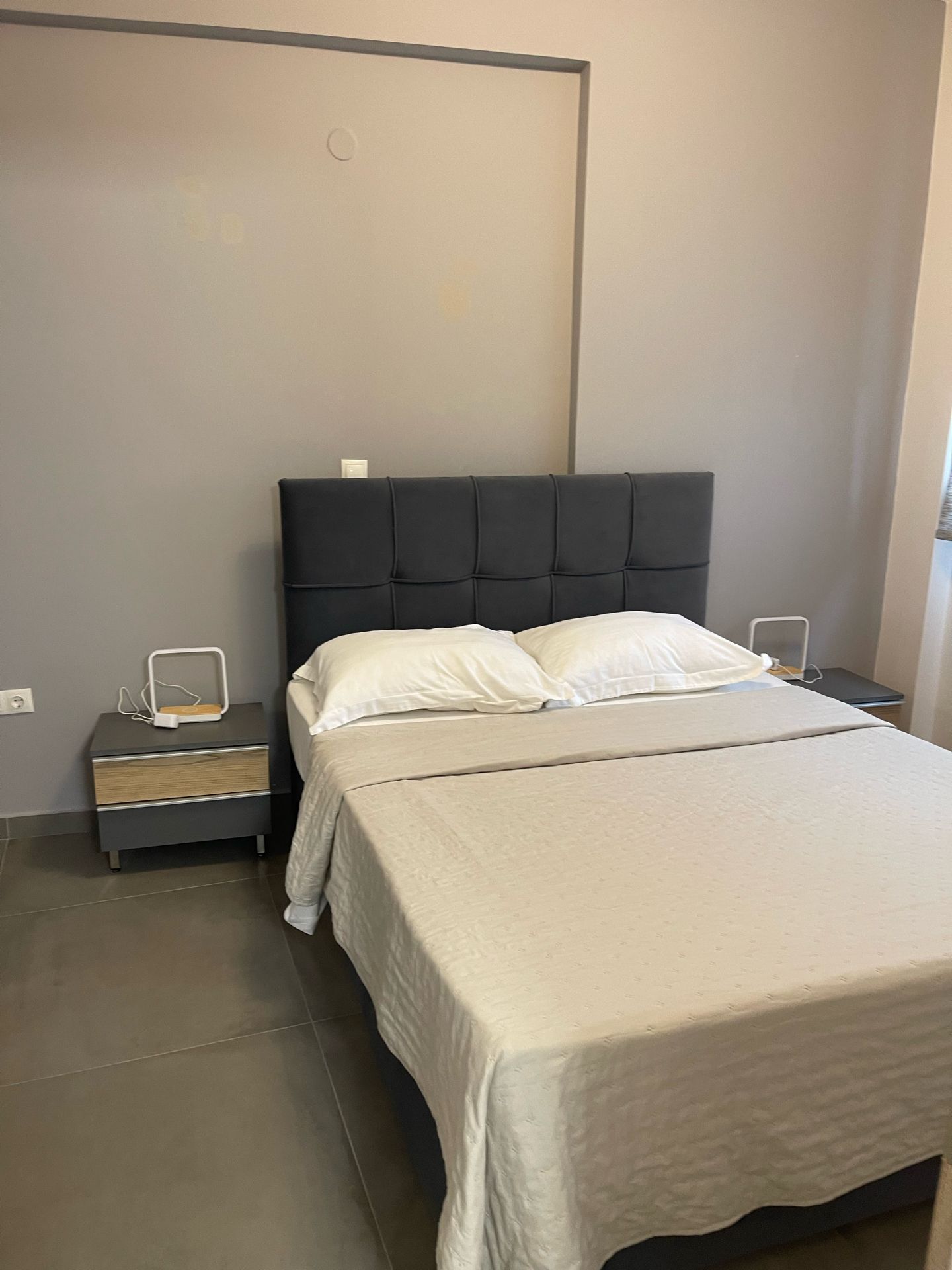 Quarto com cama, mesa de cabeceira e estação de carregamento na parede.