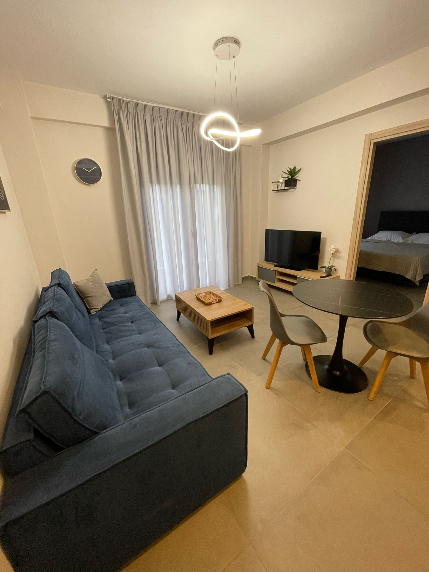 Ein modernes Wohnzimmer mit einem blauen Sofa, einem Holztisch, einem Esstisch mit Stühlen, einem Flachbildfernseher und einem Bett im Hintergrund.