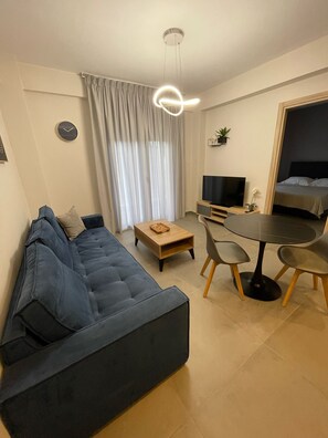 Ein modernes Wohnzimmer mit einem blauen Sofa, einem Holztisch, einem Esstisch mit Stühlen, einem Flachbildfernseher und einem Bett im Hintergrund.