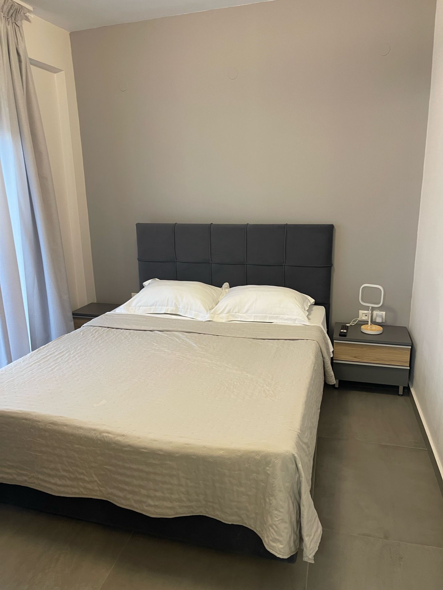 Een keurig opgemaakt bed met hoofdbord, een nachtkastje met een lamp en een gordijn voor het raam.