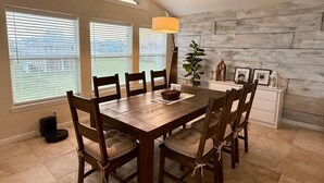 Uma sala de jantar com mesa e cadeiras de madeira, luminária pendente e vista através de grandes janelas.