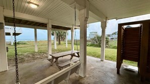 Área externa coberta com baloiço e mesa de piquenique, com vista para uma área gramada e o oceano.