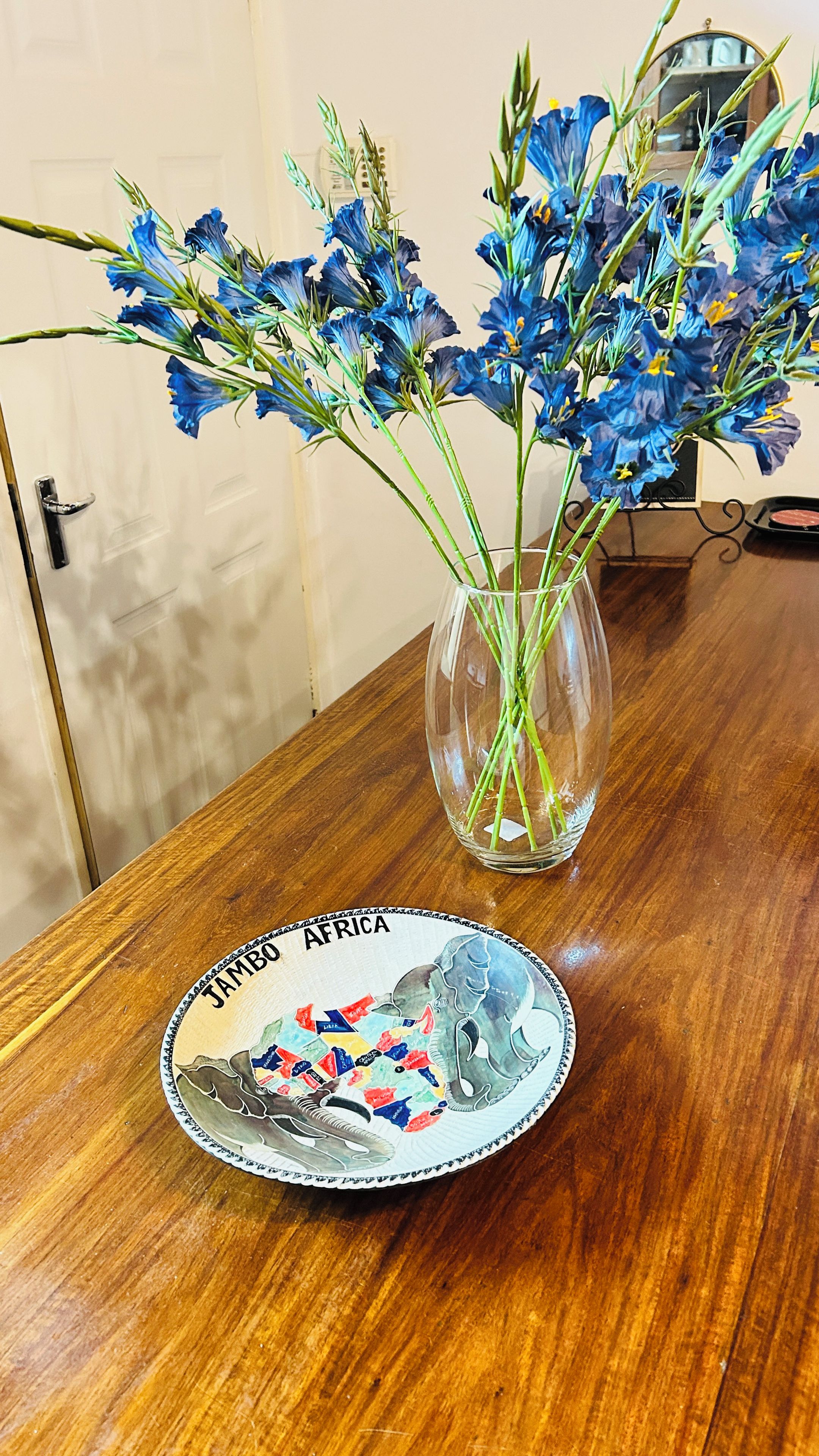 Una mesa de madera con un florero de vidrio transparente que contiene flores azules y un plato con un mapa e ilustración de un elefante.