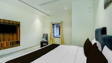 A hotel room with a bed, a TV mounted on the wall, a small table with a mirror, and a window with curtains.