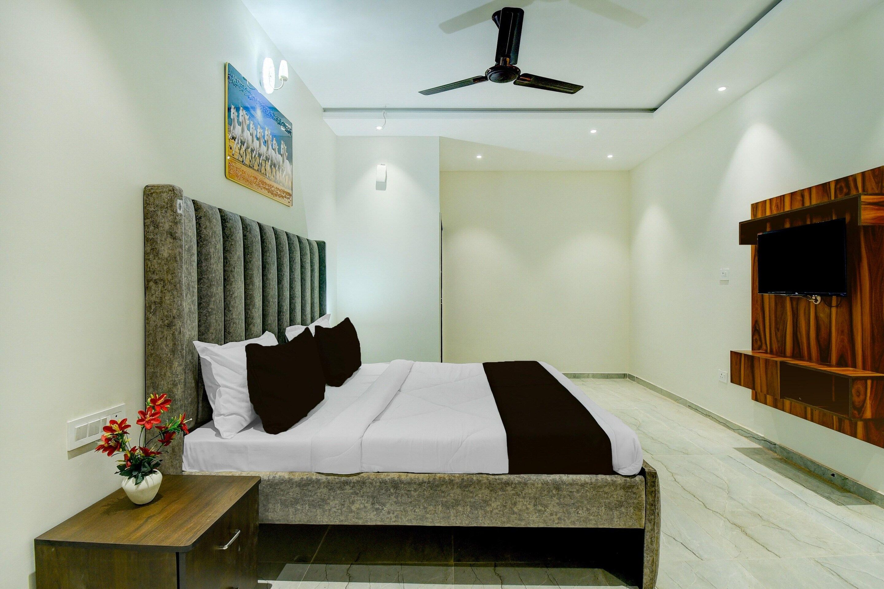 A modern hotel room with a bed, a nightstand, a vase with flowers, a ceiling fan, and a wooden panel wall with a TV.