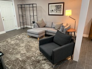 A living room with a sectional sofa, a small armchair, a floor lamp, a framed picture on the wall, and a metal shelving unit. - Private 2-bedroom apartment to rest and relax in the ancient mountains of WV! (Moorefield)
