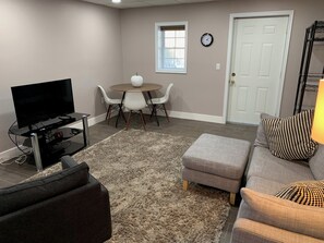 A living room with a flat-screen TV, a round dining table with chairs, a grey sofa with patterned cushions, and a white door.