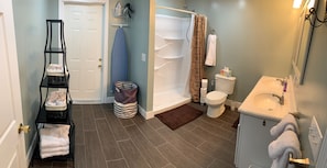 A bathroom with a shower, toilet, and sink. There is a shelf with towels and a striped trash bin. - Private 2-bedroom apartment to rest and relax in the ancient mountains of WV! (Moorefield)