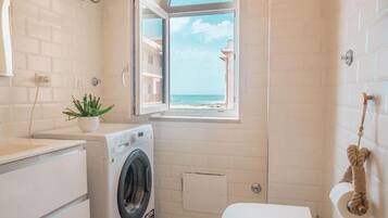 Un baño pequeño con lavadora, inodoro y ventana con vista a la playa.
