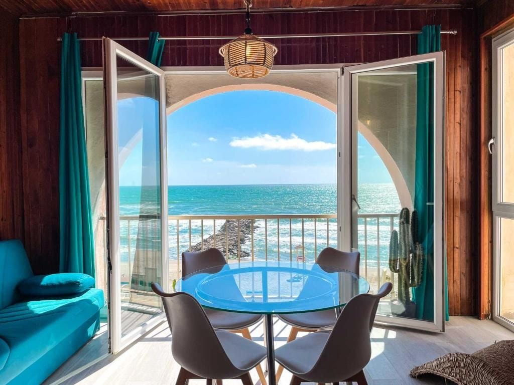 Ein Zimmer mit Glastisch und Stühlen, einer Couch und Blick auf das Meer durch eine offene Tür.