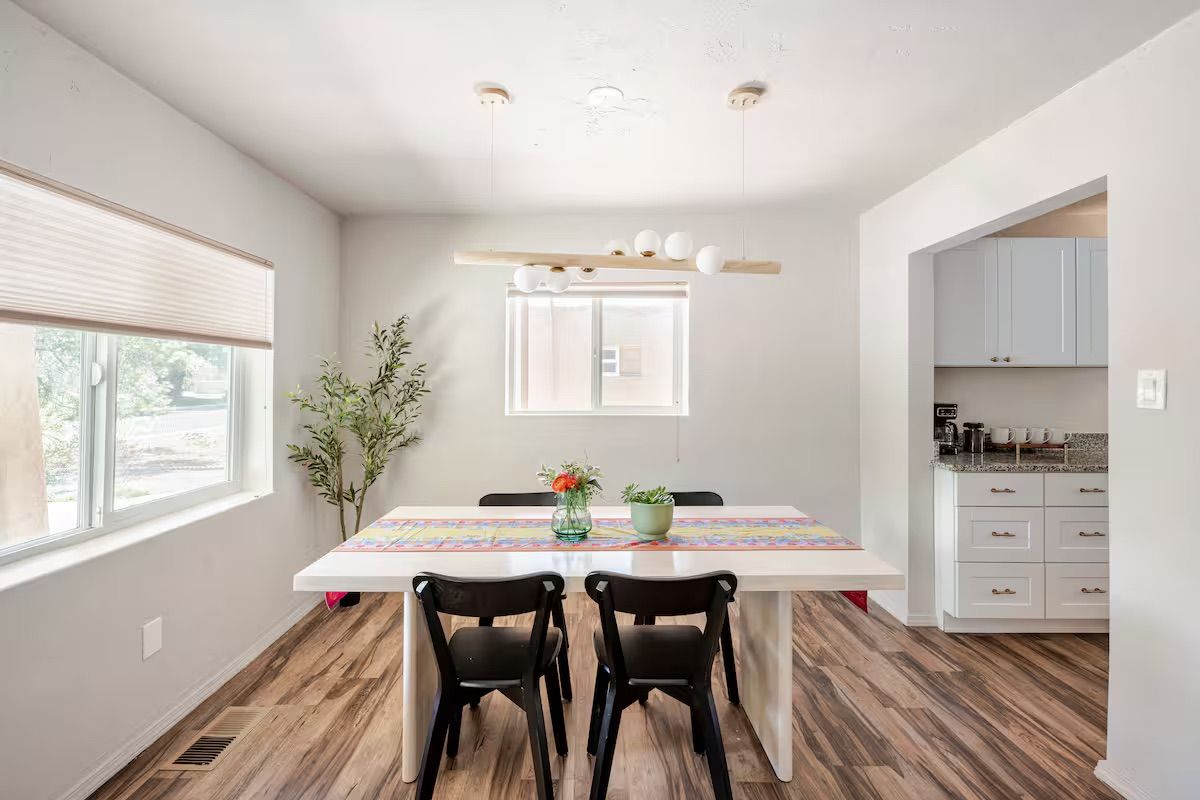 A dining area with a white table, two black chairs, a wooden floor, and a kitchen area with white cabinets.