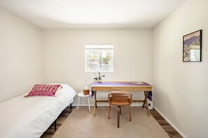 A bedroom with a bed, a desk, a chair, a window, and a wall-mounted picture. - Casa Serena (Albuquerque)