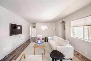 A modern living room with a flat-screen TV mounted on the wall, a white sofa, a small wooden table with a vase of flowers, and a black stool. - Casa Serena (Albuquerque)