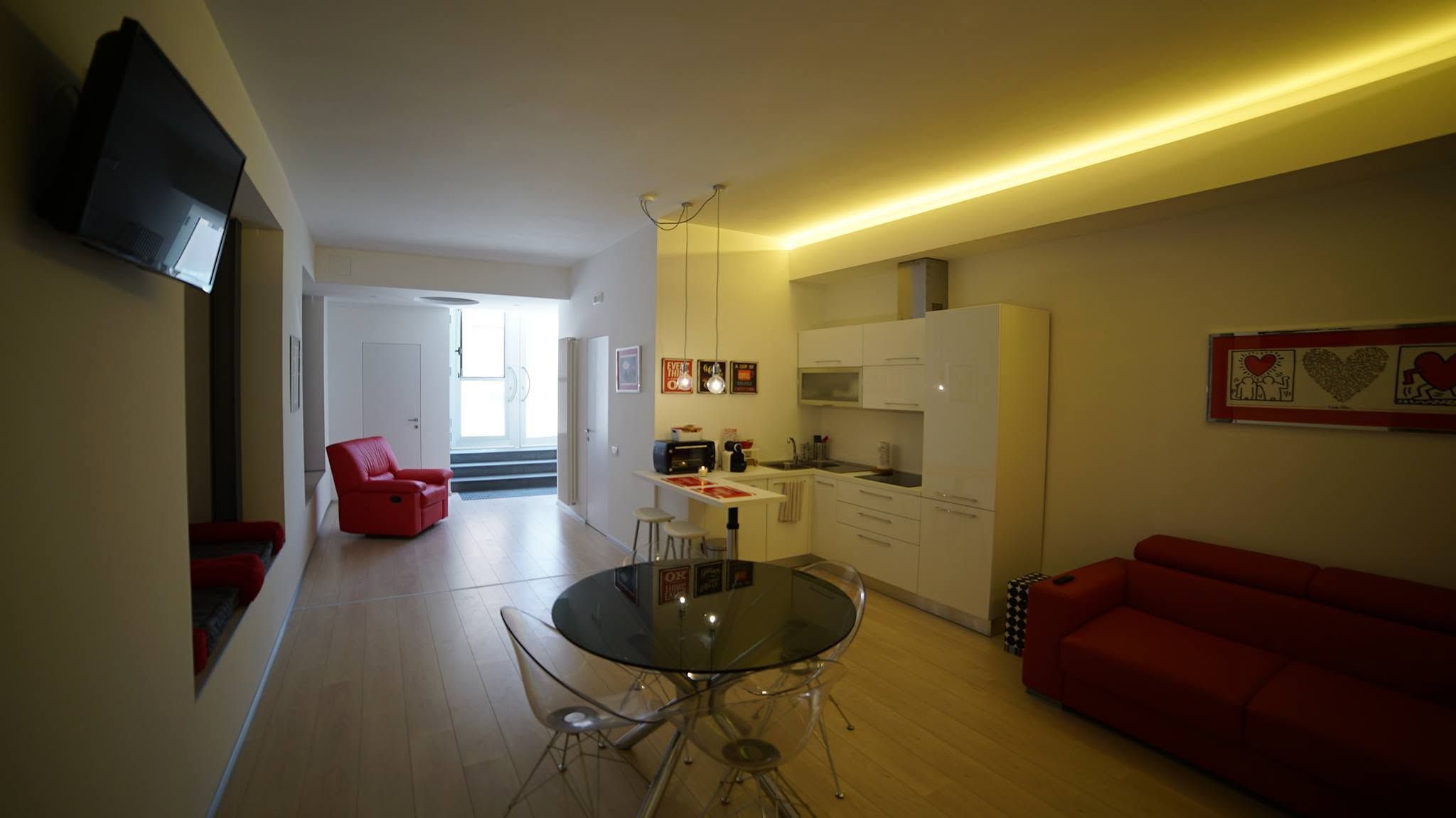 A modern living space with a glass dining table, red furniture, a kitchen area, and a flat-screen TV mounted on the wall.