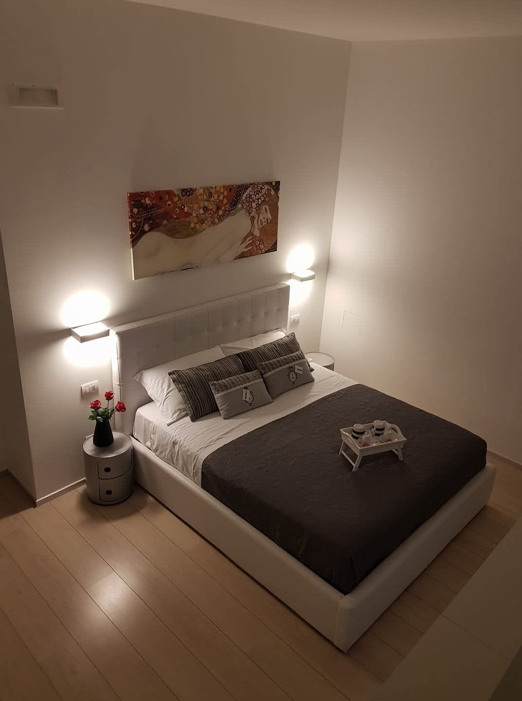 Un dormitorio moderno con cabecera blanca, un cuadro grande sobre la cama, lámparas de noche y una mesita de noche con un florero.