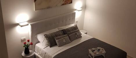 Un dormitorio moderno con cabecera blanca, un cuadro grande sobre la cama, lámparas de noche y una mesita de noche con un florero.