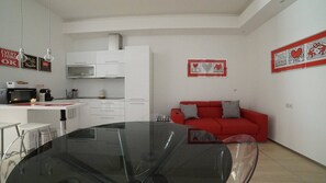 Eine moderne Küche mit weißen Schränken, einer Mikrowelle und einer Kaffeemaschine. Es gibt ein rotes Sofa mit gemusterten Kissen und ein gerahmtes Bild an der Wand.