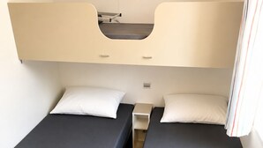 Een kamer met twee eenpersoonsbedden, elk met een kussen en een opgevouwen deken. Daartussen bevindt zich een klein nachtkastje.