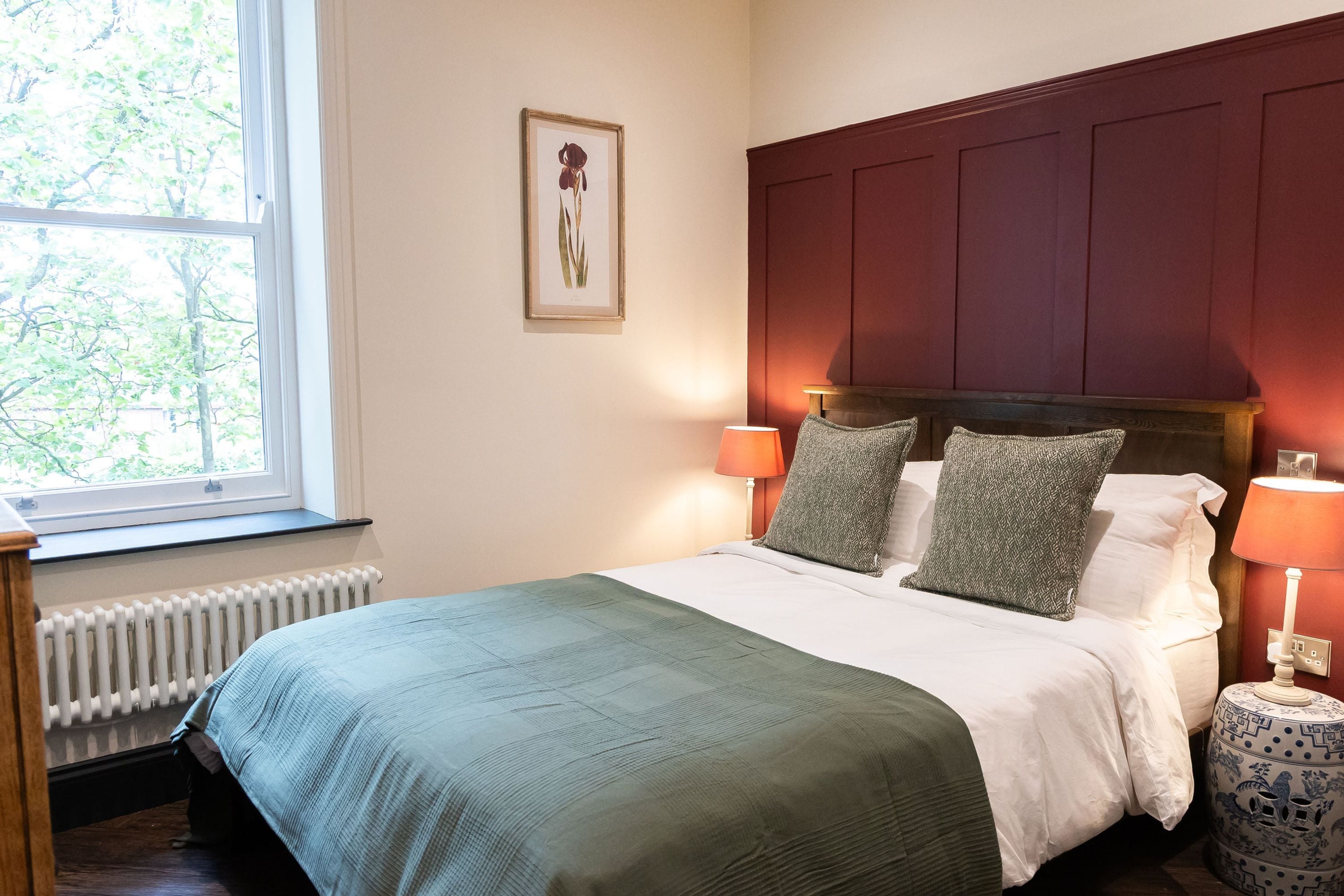 Ein ordentlich bezogenes Bett mit einer grünen Tagesdecke, zwei Kissen und einem Kopfteil. Es gibt ein Fenster mit Blick auf Bäume, eine Heizung und einen Nachttisch mit einer Lampe.