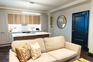 A modern living room with a beige sofa, a coffee table, and a kitchen area with wooden cabinets and a white countertop. - Summerhill in Newcastle (Newcastle)