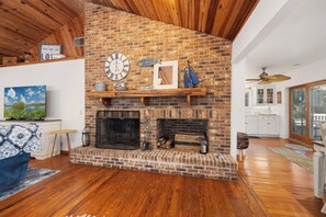 A living room with a brick fireplace, wooden beams, a clock, and a television. - Lake Santa Fe Getaway | Private Dock + Kayaks (Melrose)