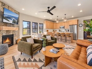 A modern living room with a fireplace, sofas, a coffee table, and a kitchen area with wooden cabinets and stainless steel appliances.