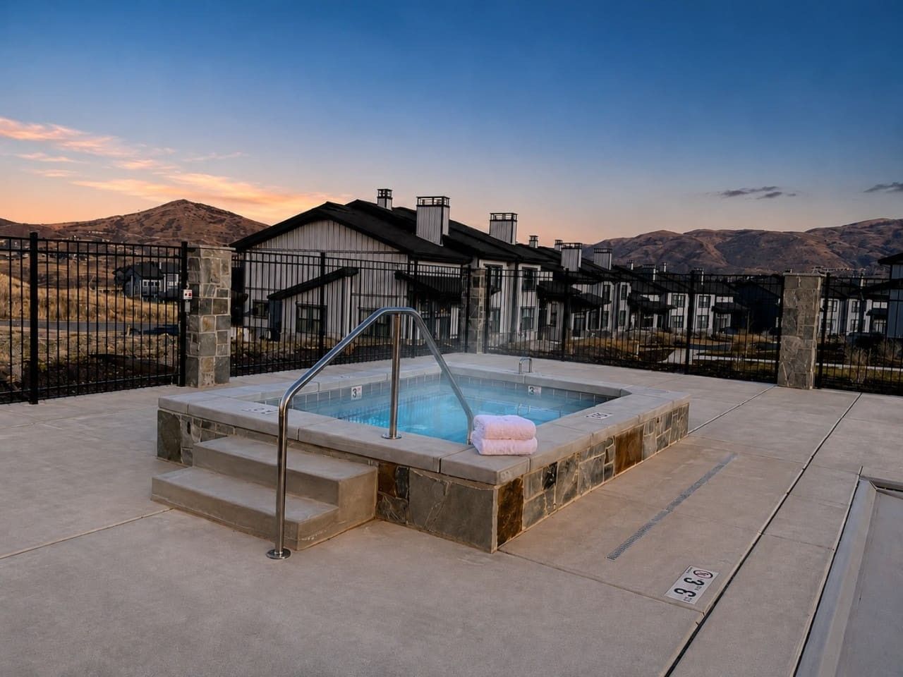 Un bain à remous doté d’une balustrade en métal, entouré d’une terrasse en pierre, avec une vue sur les montagnes et une clôture délimitant l’espace.