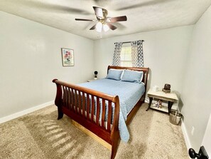 A bedroom with a bed, a nightstand, a ceiling fan, a window with curtains, and a carpeted floor. - Stillwater Cottage at Norfork Lake (Mountain Home)