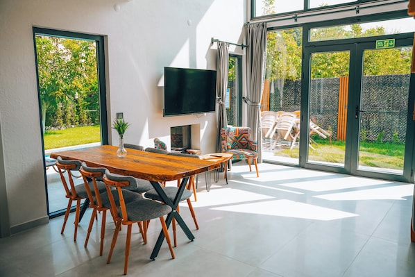 A modern dining area with a wooden table and chairs, a flat-screen TV mounted on the wall, and large sliding glass doors leading to an outdoor space.