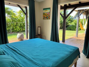 Ein Schlafzimmer mit einem großen Bett, einem Gemälde an der Wand und Blick auf einen Garten durch offene Vorhänge.