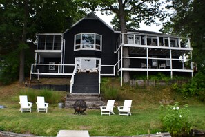 A two-story house with a balcony, a front porch with chairs, and a small table. - 5BR All-Sports Lakehouse · Hot Tub + Pontoon · Portage (Portage)