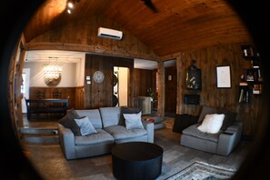 A cozy living room with a large gray sofa, a smaller armchair, and a coffee table. There is a wooden ceiling and a wall-mounted air conditioner. - 5BR All-Sports Lakehouse · Hot Tub + Pontoon · Portage (Portage)