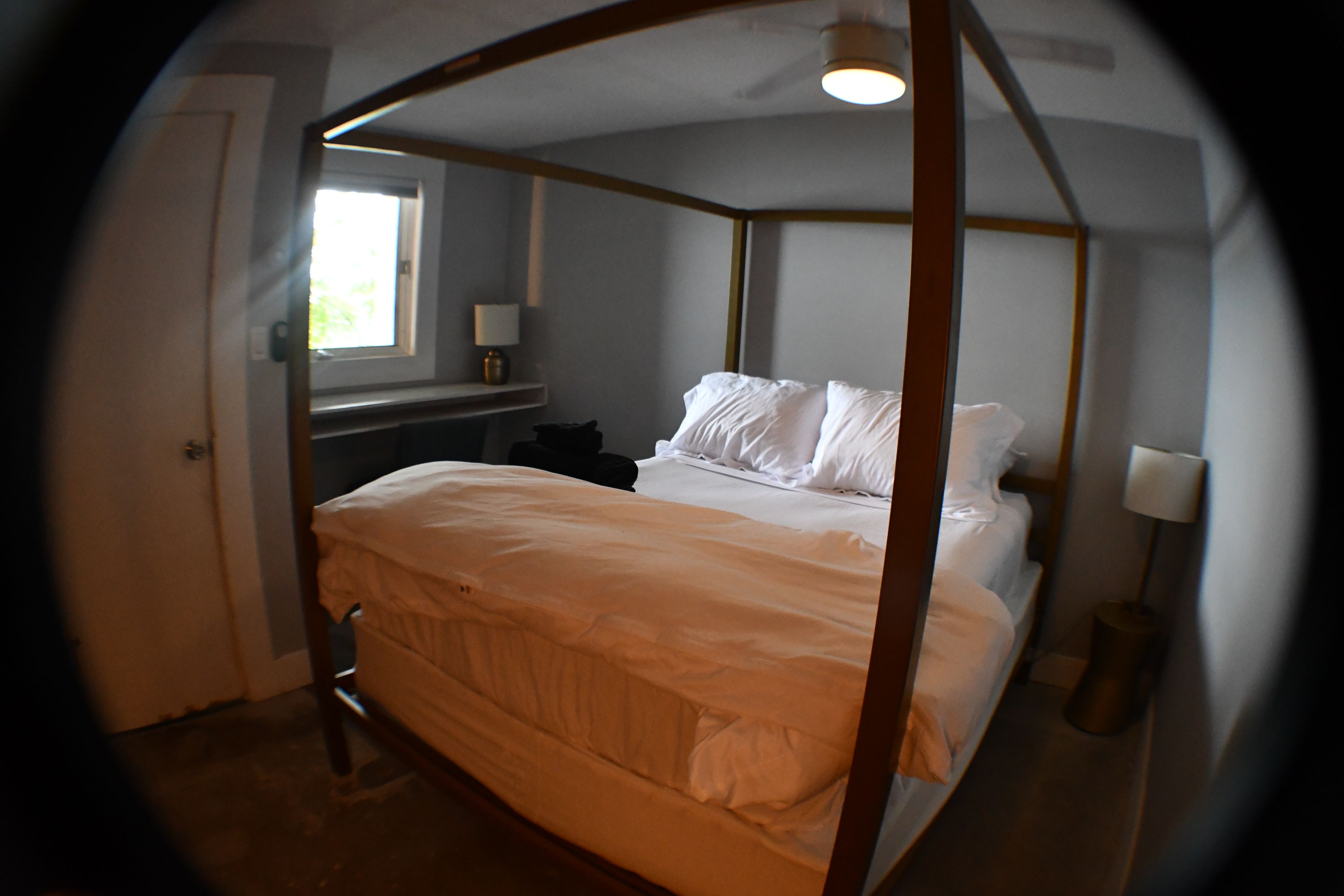 A bedroom with a four-poster bed, a bedside table, a lamp, and a window.