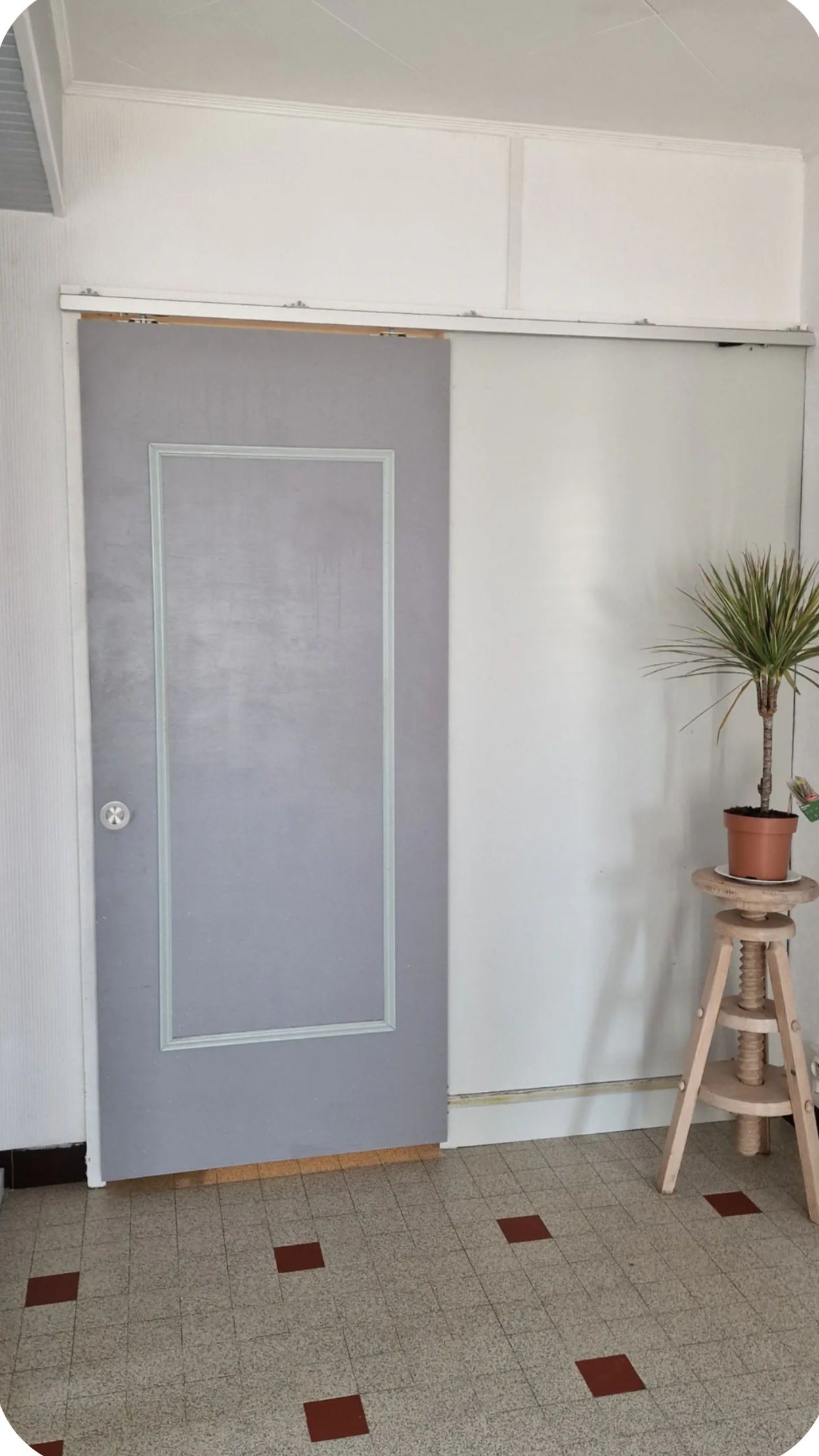 Een kamer met een grijze deur, een potplant op een houten kruk, en een betegelde vloer met rode vierkanten.