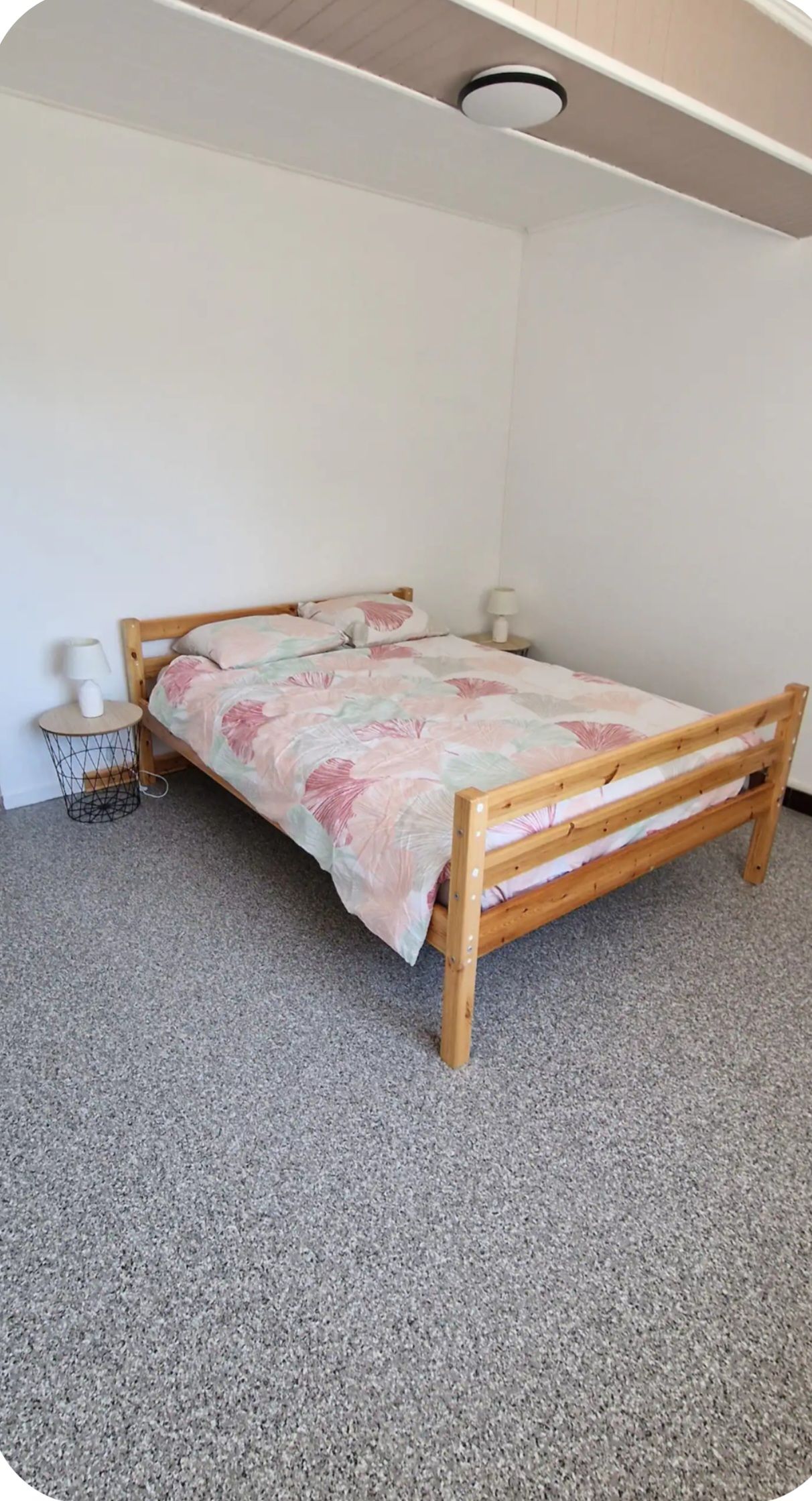 A single bed with a wooden frame and a floral bedspread in a room with a plain white wall and a ceiling light fixture.