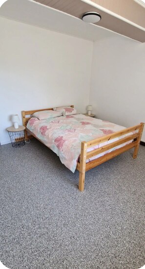 A single bed with a wooden frame and a floral bedspread in a room with a plain white wall and a ceiling light fixture.