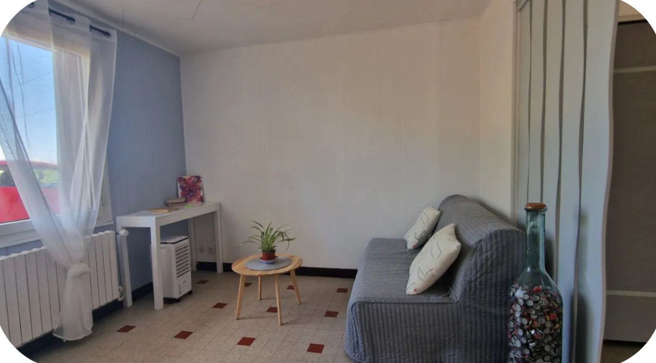 Ein Zimmer mit einer grauen Couch, einem kleinen runden Tisch mit einer Pflanze, einem Heizkörper und einem Fenster mit transparenten Vorhängen.