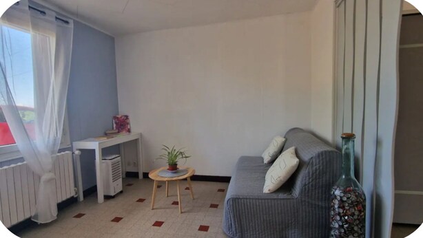 Una habitación con un sofá gris, una mesita redonda con una planta, un radiador y una ventana con cortinas translúcidas.