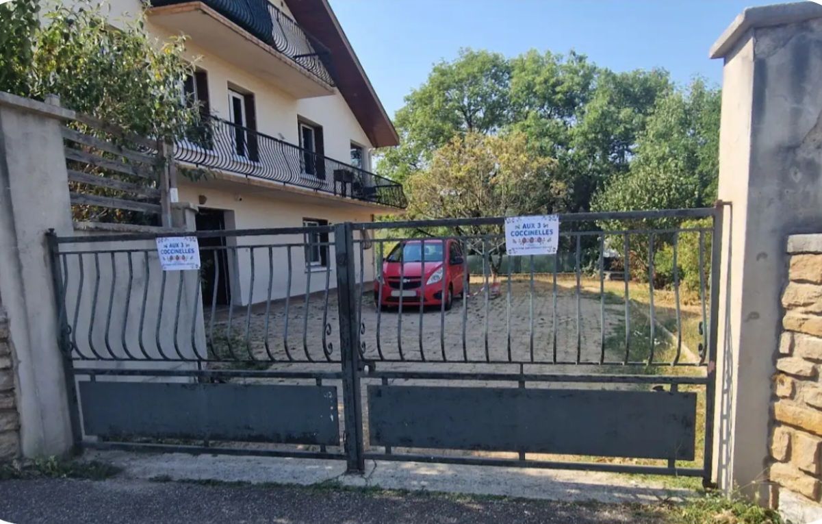 Une propriété privée, fermée par un portail, où se trouve une voiture rouge garée à l’intérieur, ainsi qu’un balcon et une enseigne indiquant la présence d’un restaurant.