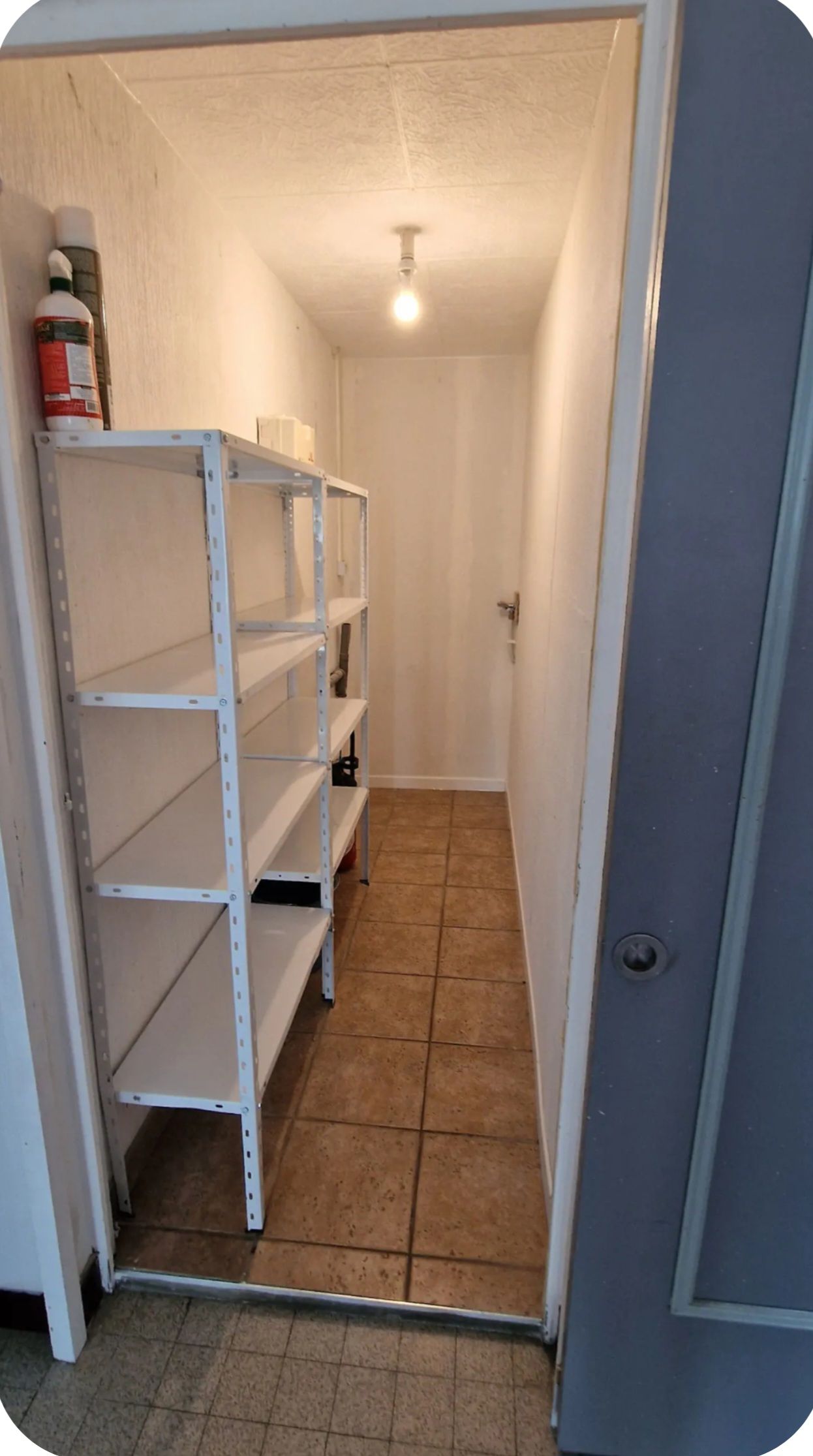 Un couloir étroit, avec une étagère métallique adossée au mur, un sol carrelé et une porte au fond.