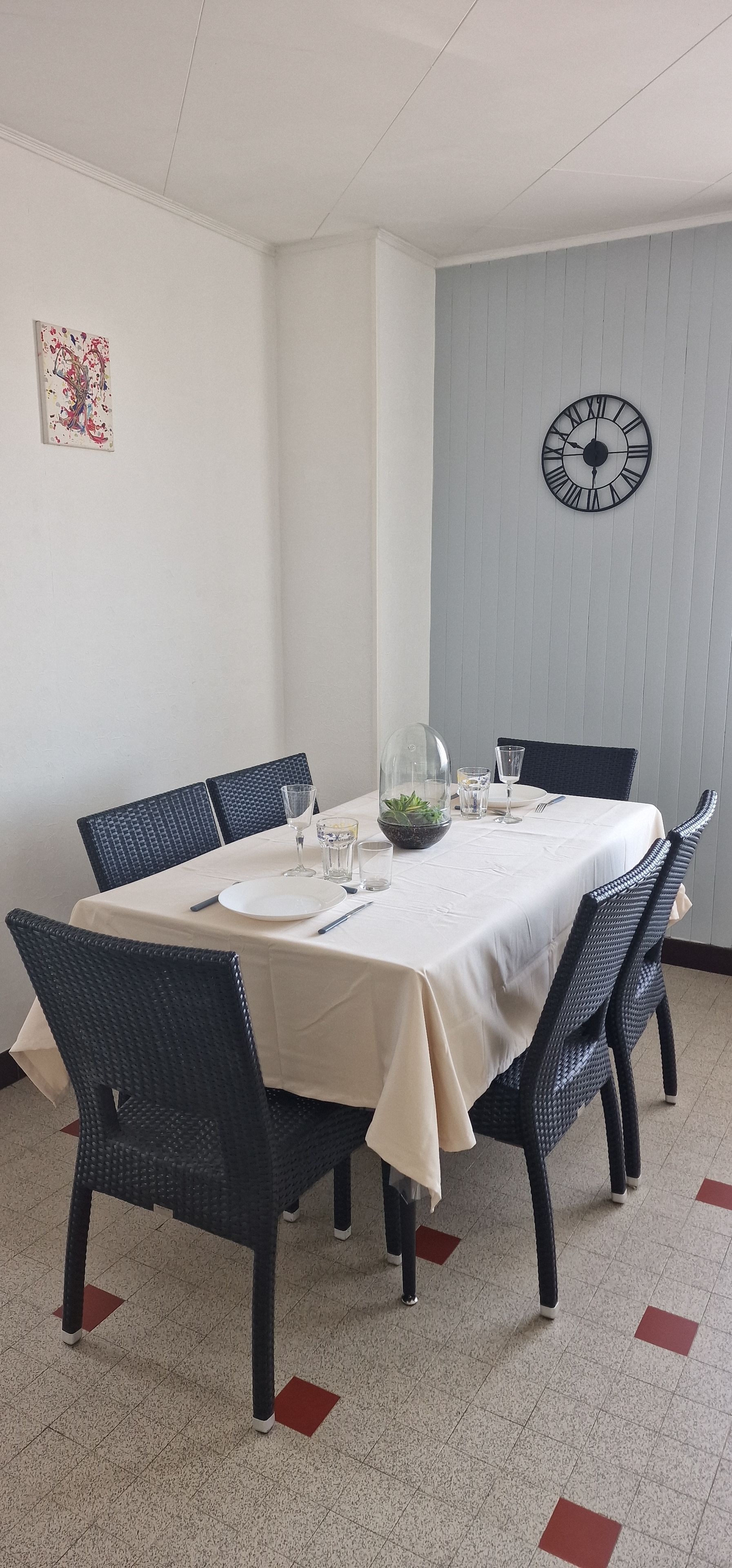 Ein Essbereich mit einem runden Tisch, gedeckt für vier Personen, umgeben von Korbstühlen, einer Uhr an der Wand und einer kleinen Pflanze auf dem Tisch.