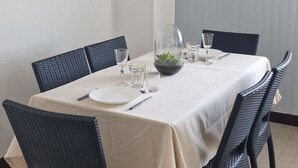 Un comedor con una mesa redonda dispuesta para cuatro personas, rodeada de sillas de mimbre, un reloj en la pared y una planta pequeña sobre la mesa.