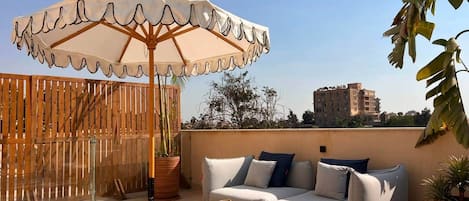 A rooftop terrace with a sofa, a table, an umbrella, and a wooden fence.