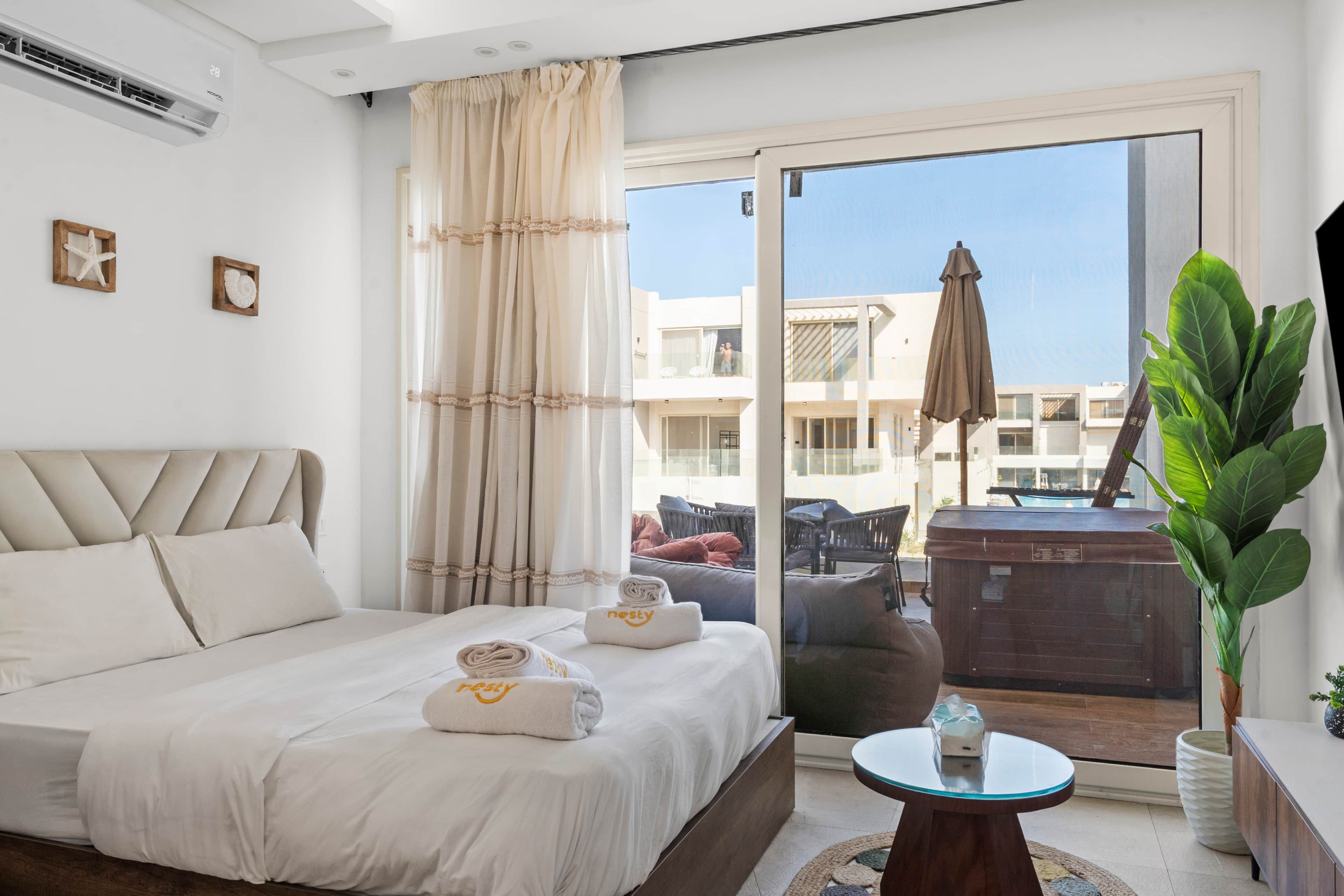 Habitación de hotel con cama, una mesa redonda, una planta en maceta y un balcón con mobiliario exterior.