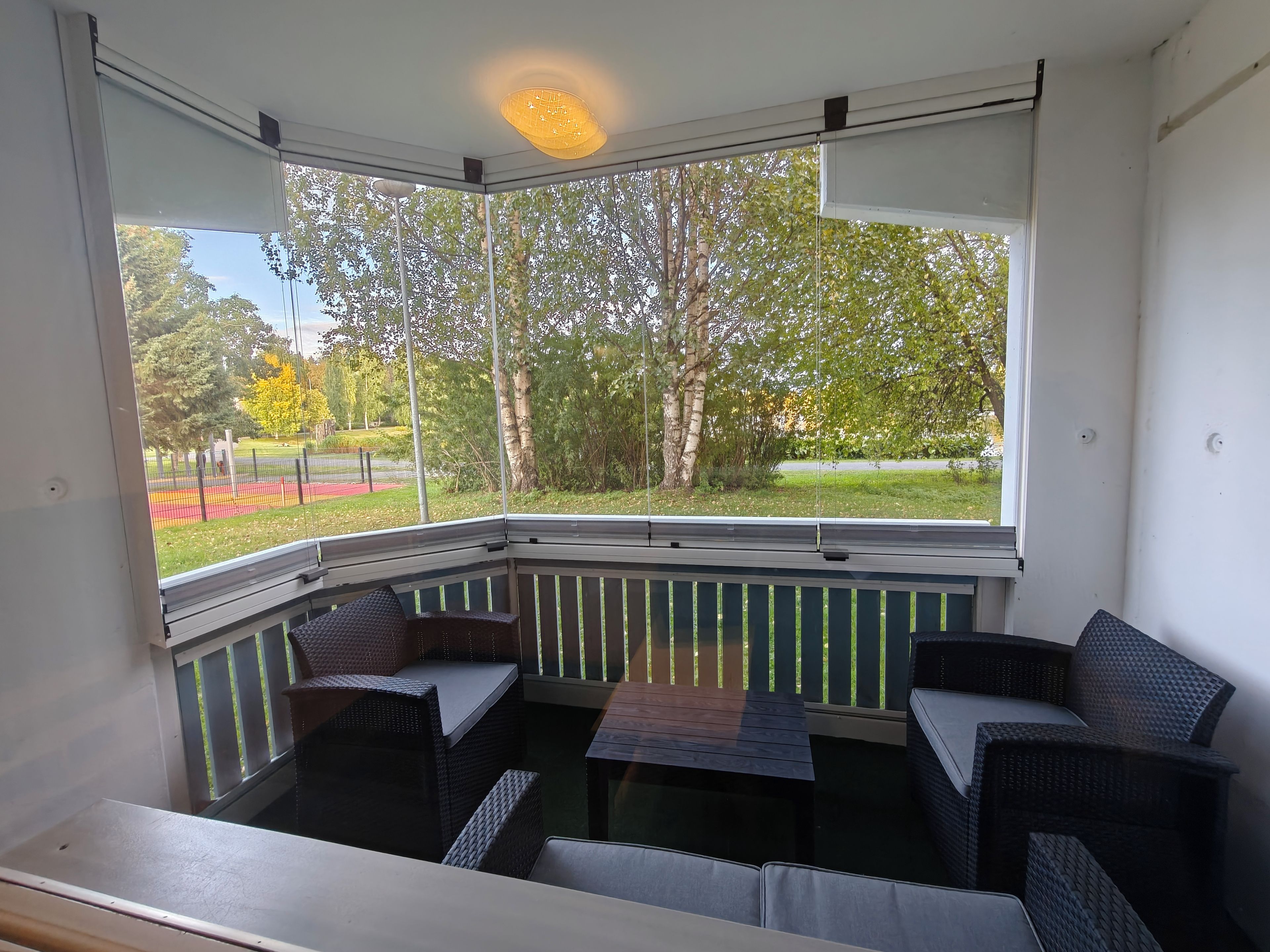 Un balcon d’angle avec des meubles en osier, une vue sur un parc et un luminaire au plafond.