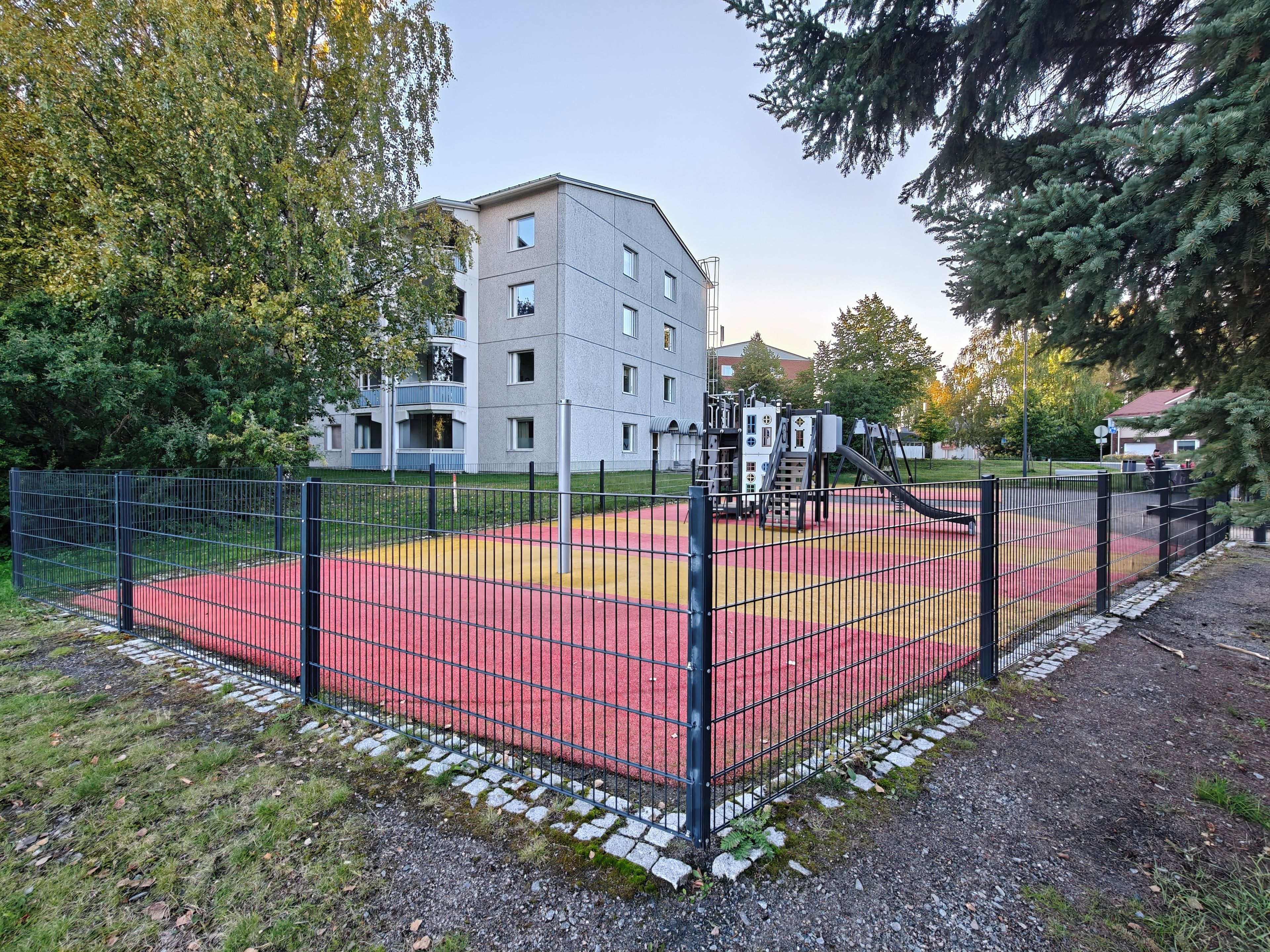 Une aire de jeux clôturée, avec des surfaces rouges et jaunes, entourée d’arbres et bordée par un bâtiment en arrière-plan.