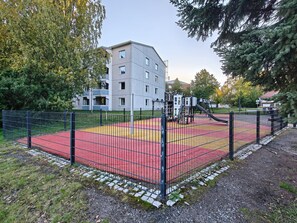 Ein von einer Zähe umfasster Spielbereich mit roten und gelben Belägen, umgeben von Bäumen und einem Gebäude im Hintergrund.