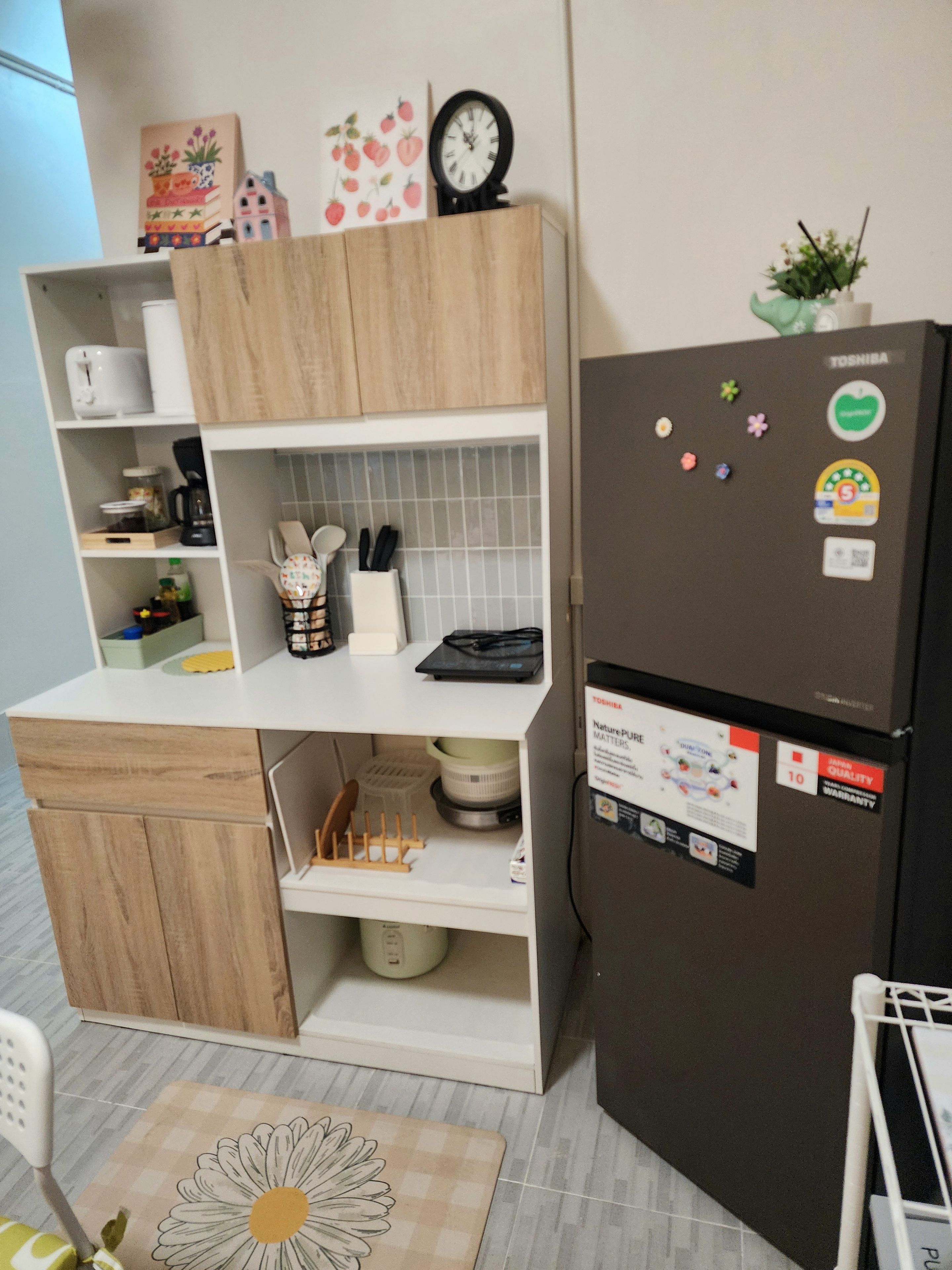A compact kitchen with wooden cabinets, a white countertop, a refrigerator with magnets, and a small clock on the wall.